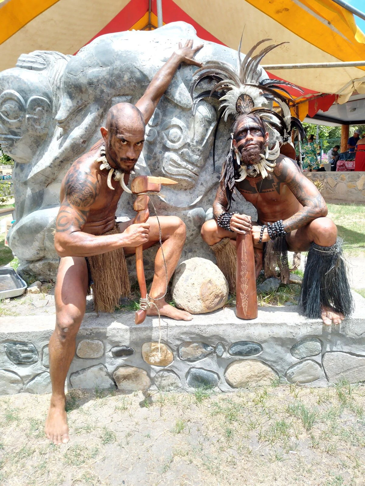 Two tattooed Marquesan warriors in traditional dress, with feather headdresses and wooden weapons, in front of a stone carving
