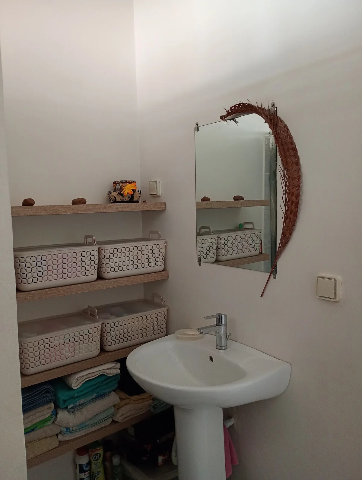 Washbasin under a mirror decorated with a woven palm frond, next to shelves with baskets and towels