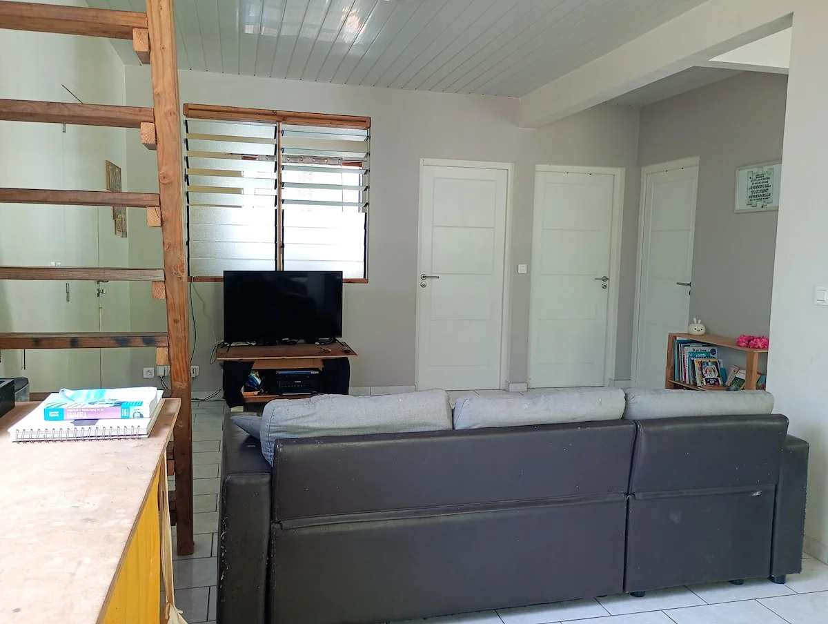Living room with a grey sofa, a TV and a wooden staircase, in front of louvred windows
